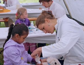 Man sieht eine Frau im Doktorkittel, die einen Teddy untersucht. Ein kleines Mädchen sieht ihr interessiert zu.