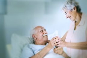 Man sieht eine alte Dame die einem älteren Mann ein Glas Wasser reicht